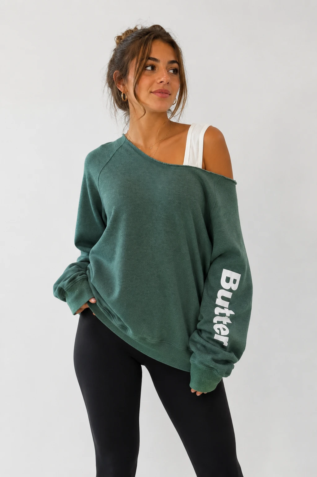 Dark forest green off-the-shoulder sweatshirt with white 'Butter' text printed vertically on the left sleeve, worn by a model with black leggings.