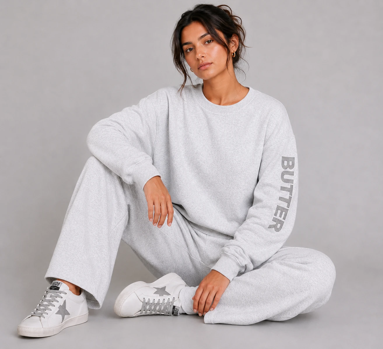 Model sitting on floor in a light grey heather off-the-shoulder set. Rhinestone logo is visible on the sleeve.