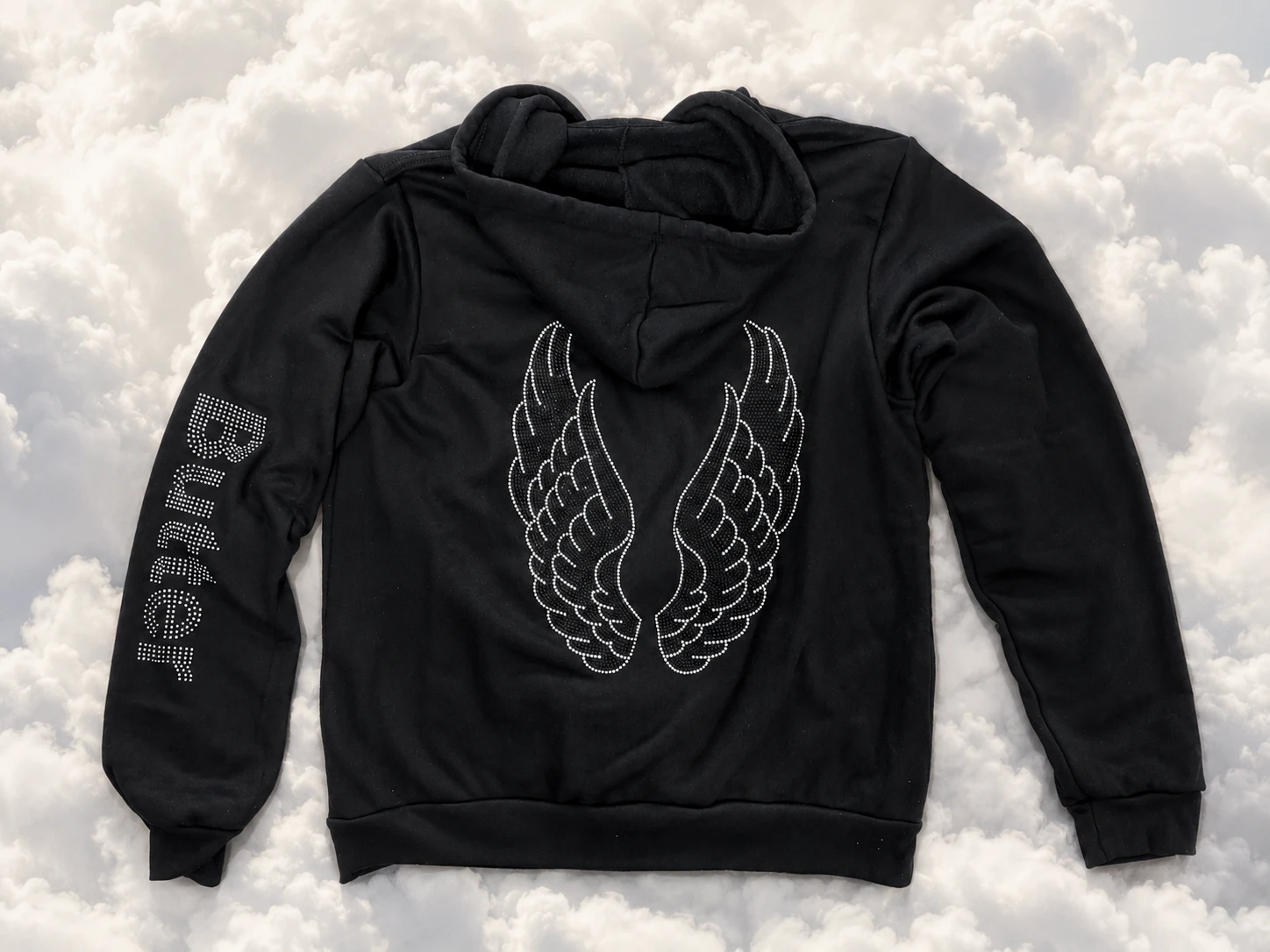 Flat lay view of the back of a black zip-up hoodie with tonal rhinestone wings and 'Butter' branding on the sleeve, placed against a cloud-filled sky background.