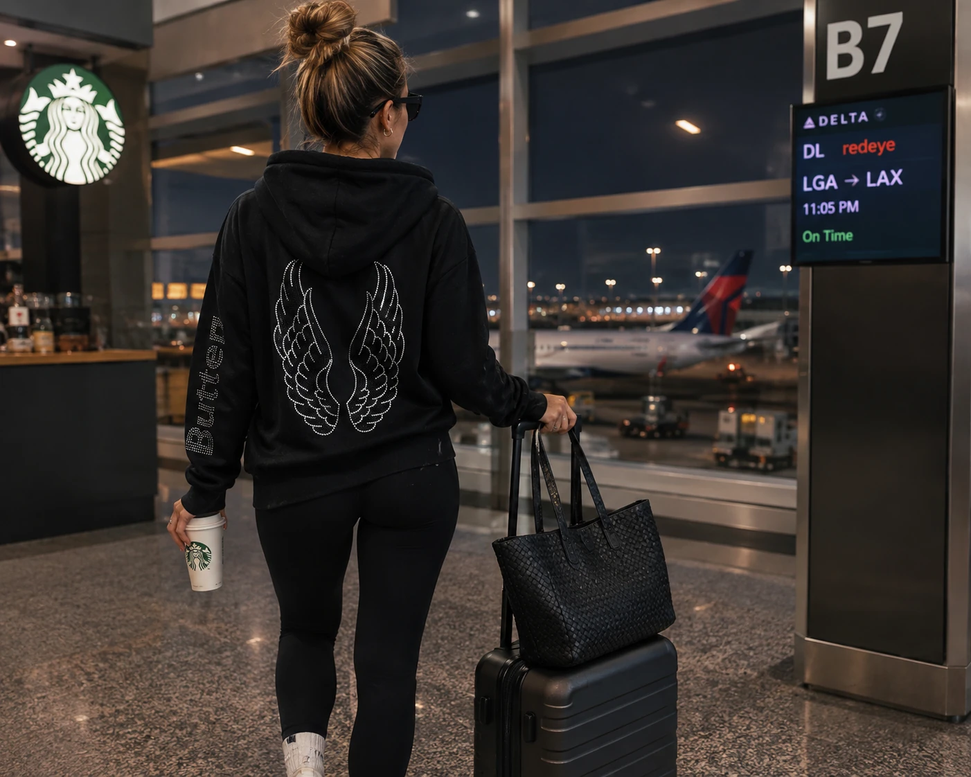 Lifestyle image of a woman in an airport wearing the black zip-up hoodie with the tonal onyx wing design on the back.
