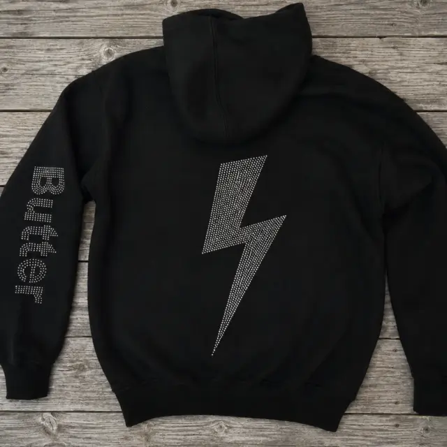 Black zip-up hoodie featuring a rhinestone lightning bolt on the back and 'Butter' text on the sleeve, displayed on a wooden surface.