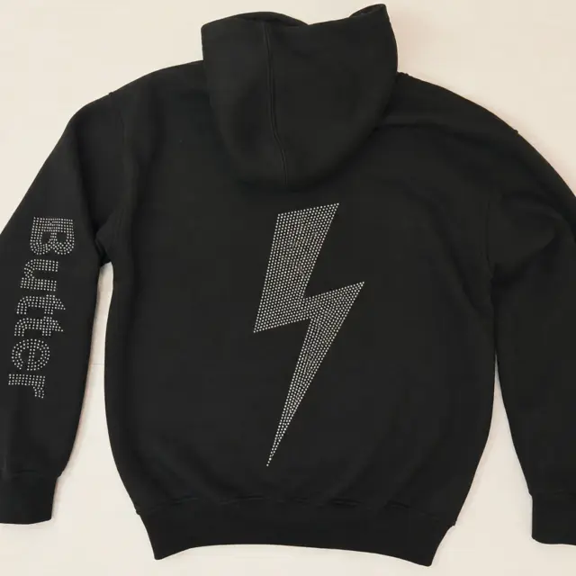 Back flat-lay of a black zip hoodie with a rhinestone lightning bolt design and 'Butter' branding on the left sleeve.