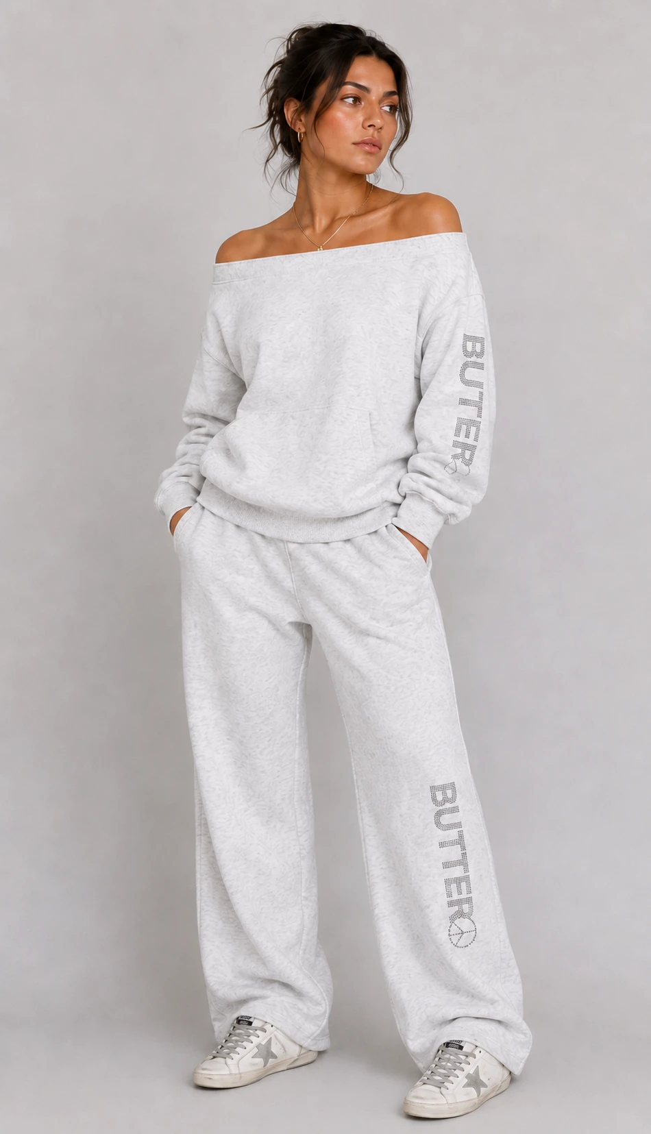 Model wearing a light grey heathered off-the-shoulder sweatshirt and matching pants set with rhinestone details.
