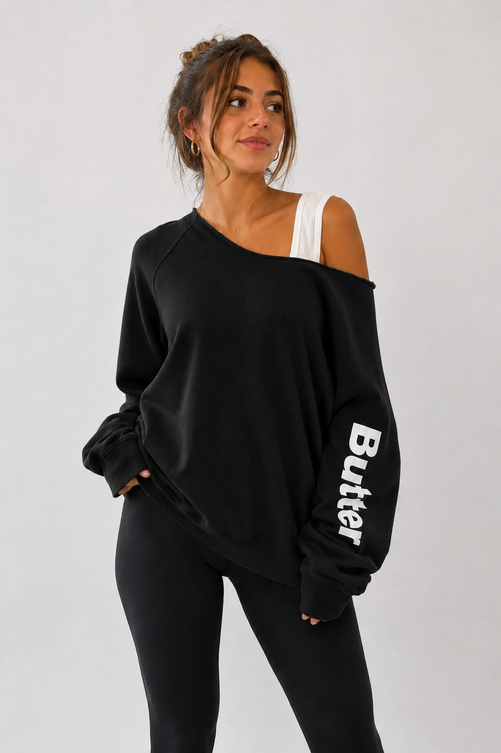 Black off-the-shoulder sweatshirt with white 'Butter' text printed vertically on the left sleeve, worn by a model with black leggings.