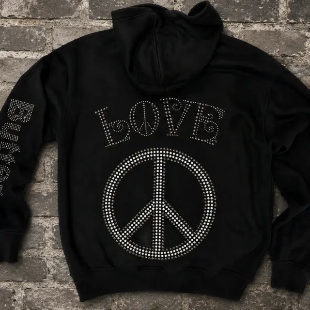 Back flat-lay of a black zip hoodie featuring rhinestone 'LOVE' text above a rhinestone peace sign on a brick background.
