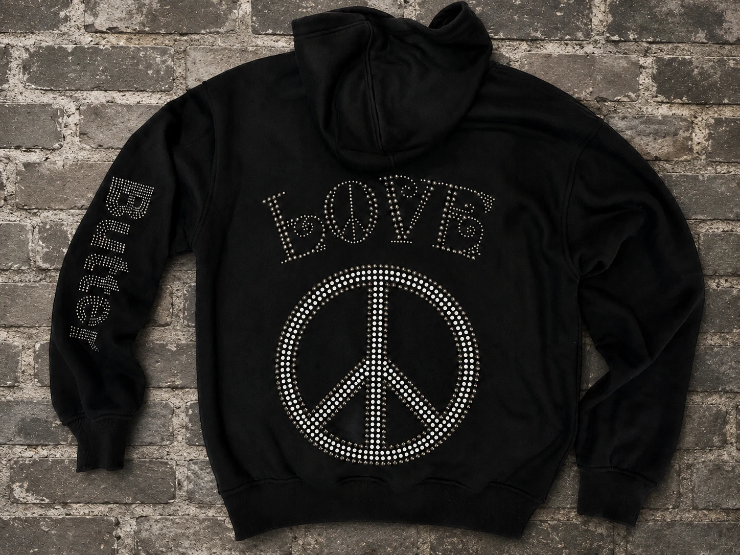 Back flat-lay of a black zip hoodie featuring rhinestone 'LOVE' text above a rhinestone peace sign on a brick background.